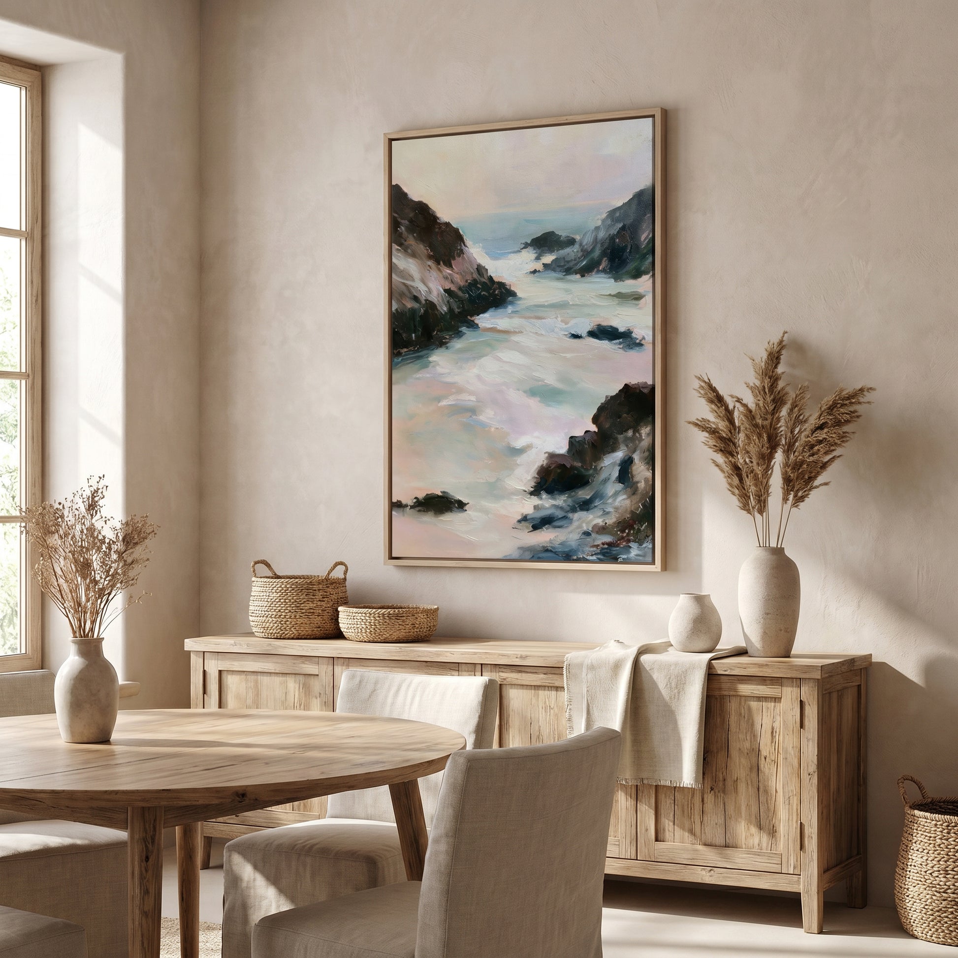 a serene dining room with a large painting of a rocky coastline hanging on the wall above a wooden table and chairs. The room has a neutral color palette, with a large window providing natural light and a view of the outdoors. The dining table is set with a vase of dried flowers and a woven basket, adding a touch of nature and rustic charm to the space.