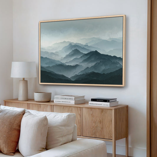 a living room with a large framed landscape painting hanging on the wall above a wooden cabinet. The painting depicts a mountainous scene with a hazy, misty atmosphere. The room also includes a white sofa, a lamp, and a stack of books on the cabinet.