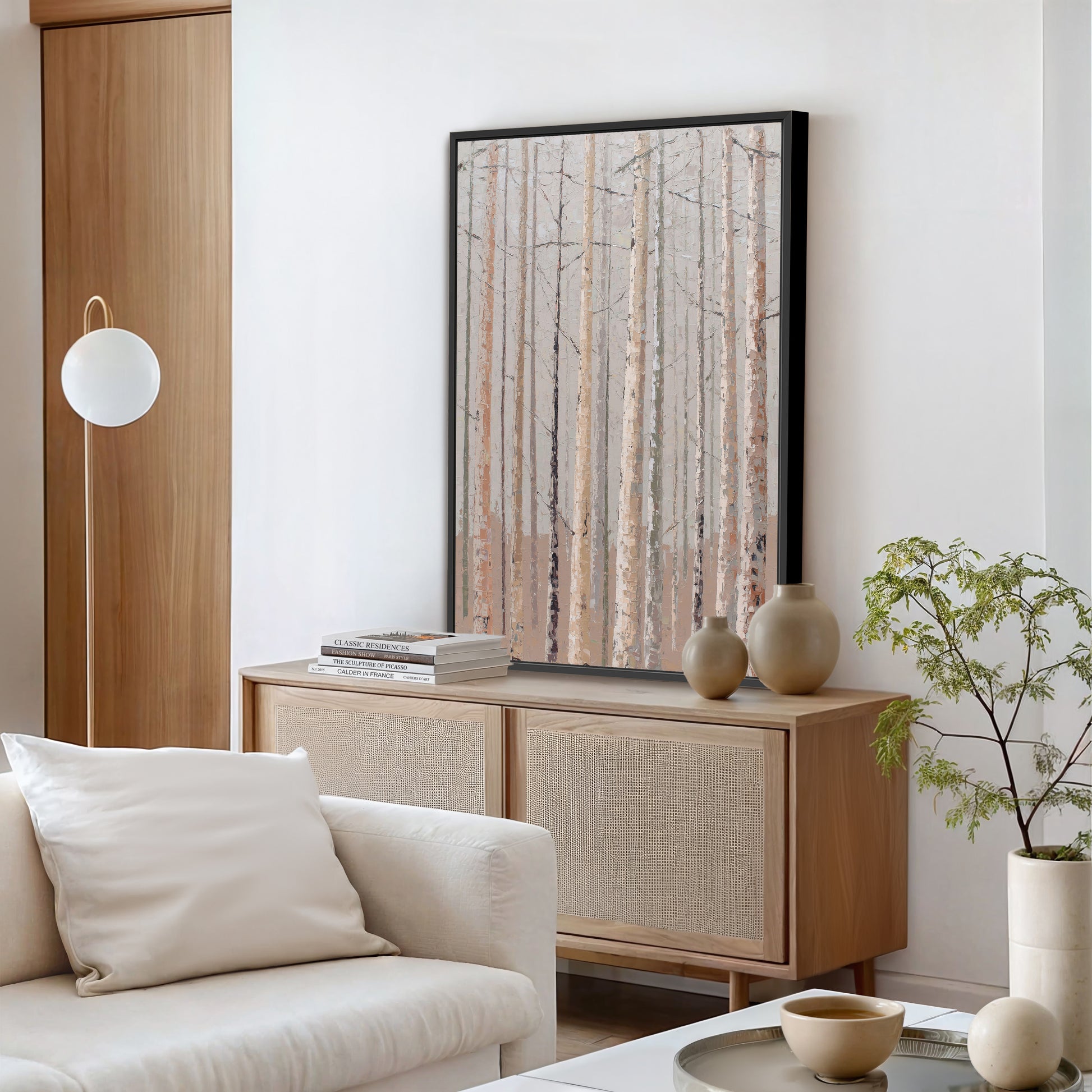 a modern living room with a large framed picture of a forest scene hanging on the wall above a wooden cabinet. The room is furnished with a white sofa, a coffee table, and a potted plant, creating a serene and inviting atmosphere.