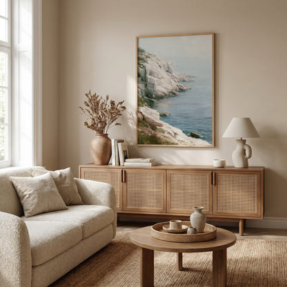 a serene living room with a large framed picture of a cliff overlooking the ocean hanging on the wall. The room features a beige sofa, a wooden sideboard, and a round coffee table with a vase and a book on it. The overall decor has a minimalist and coastal theme.