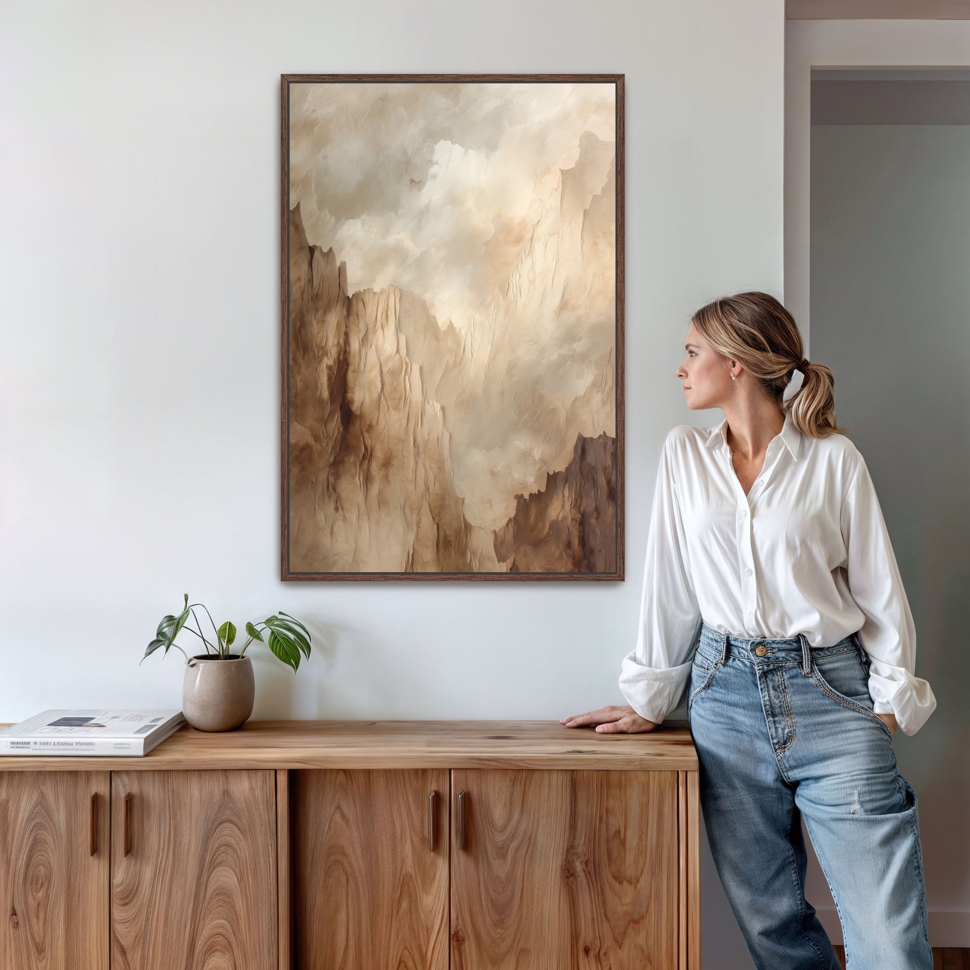 A woman in a white shirt and blue jeans stands next to a wooden cabinet, leaning against it and looking to the side. Above her, a framed painting of a landscape with a cloudy sky hangs on the wall.