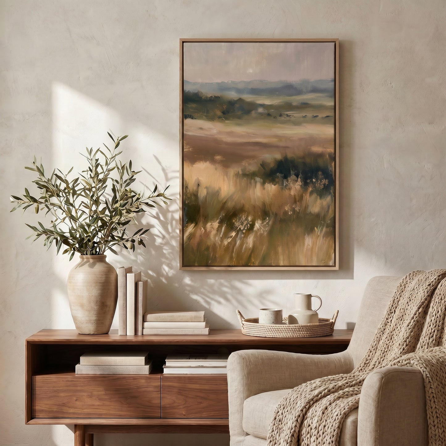 a serene living room with a large landscape painting hanging on the wall, a wooden side table with a vase of greenery and books, and a beige armchair with a cozy blanket draped over it.