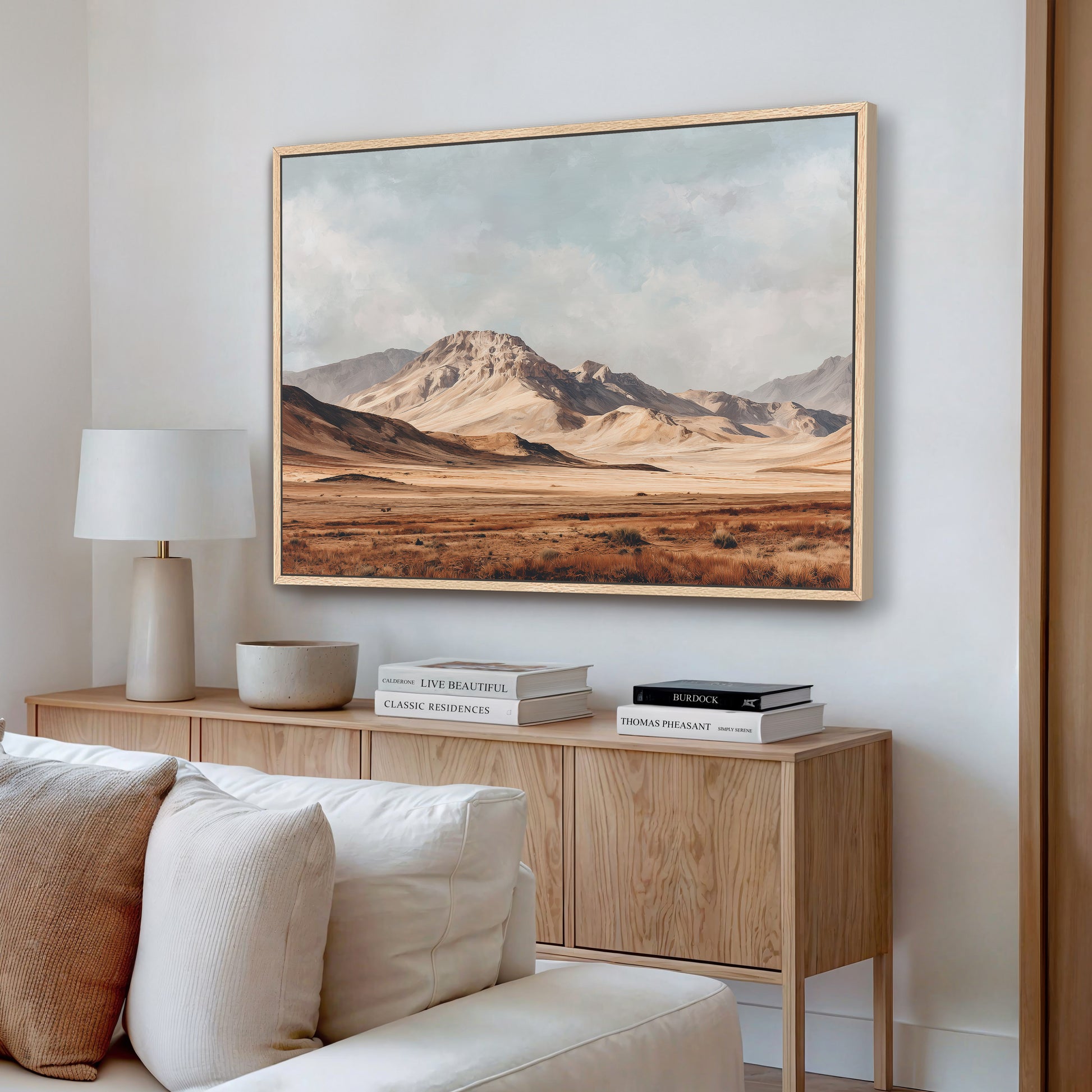 a living room with a large framed landscape painting hanging on the wall above a wooden cabinet. The painting depicts a desert scene with mountains in the background. The cabinet has a white lamp and a stack of books on top. The couch in the room is white and has a brown throw pillow.