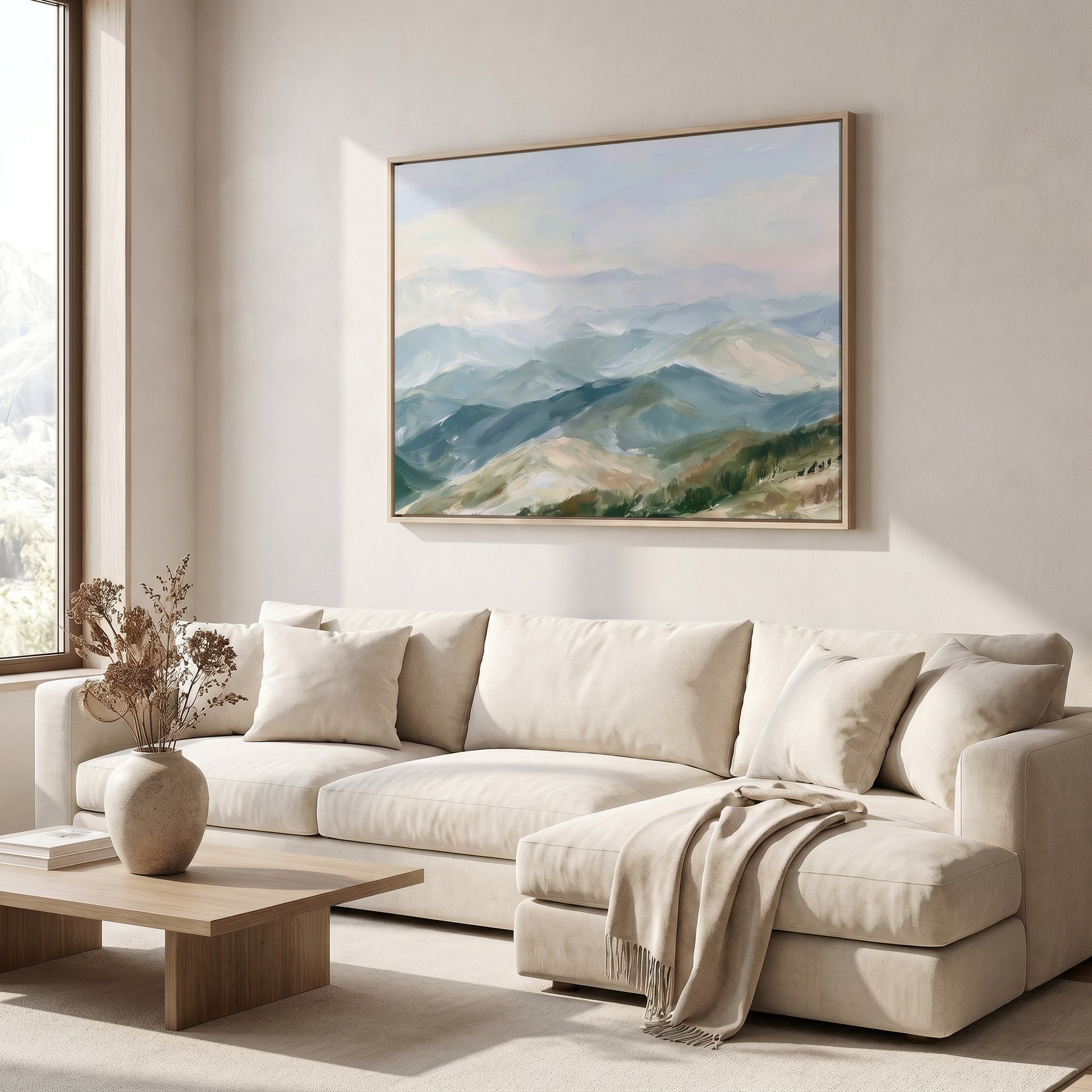 a serene living room with a large painting of a mountainous landscape hanging on the wall above a beige sofa. The sofa is adorned with a vase of dried flowers and a throw blanket, adding a touch of warmth and coziness to the space. The room is illuminated by natural light coming through a window, creating a bright and inviting atmosphere.