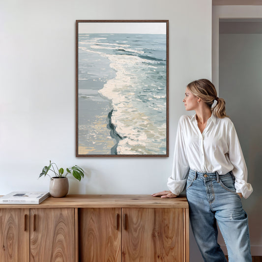 A woman in a white blouse and blue jeans stands next to a wooden cabinet, leaning against it and looking to the side. Above her, a framed painting of a serene ocean scene hangs on the wall.