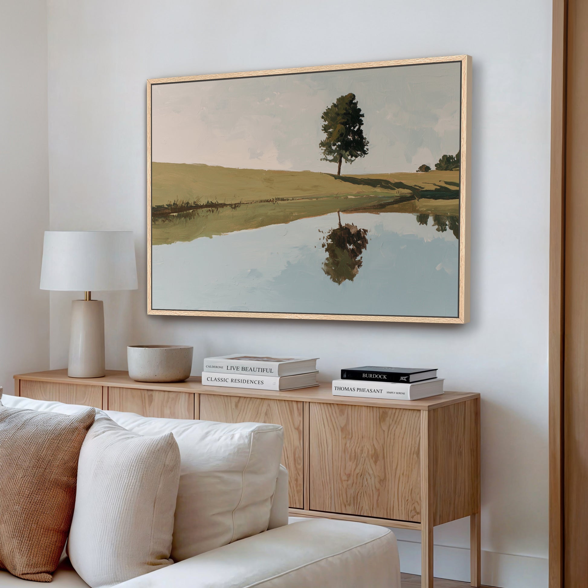 a living room with a large painting of a tree reflected in a lake on the wall. The painting is framed in a wooden frame and hangs above a wooden cabinet or console table. The cabinet has a lamp and some books on it. The room also includes a white couch and a wooden floor.