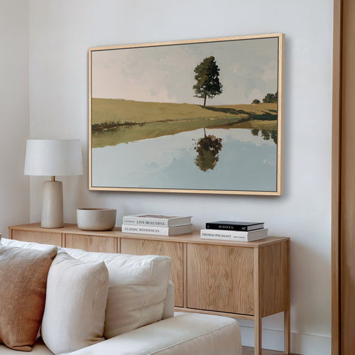 a living room with a large painting of a tree reflected in a lake on the wall. The painting is framed in a wooden frame and hangs above a wooden cabinet or console table. The cabinet has a lamp and some books on it. The room also includes a white couch and a wooden floor.