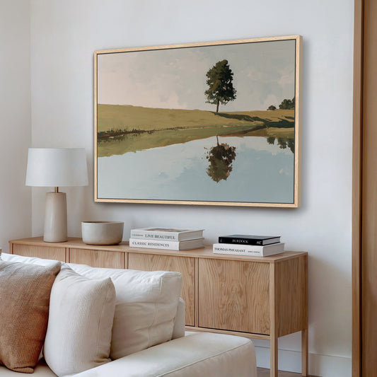a living room with a large painting of a tree reflected in a lake on the wall. The painting is framed in a wooden frame and hangs above a wooden cabinet or console table. The cabinet has a lamp and some books on it. The room also includes a white couch and a wooden floor.