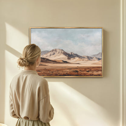 A woman stands in front of a framed painting depicting a desert landscape with mountains in the background.