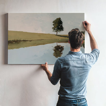 A man is hanging a painting on a wall, which depicts a serene landscape with a tree and a body of water.