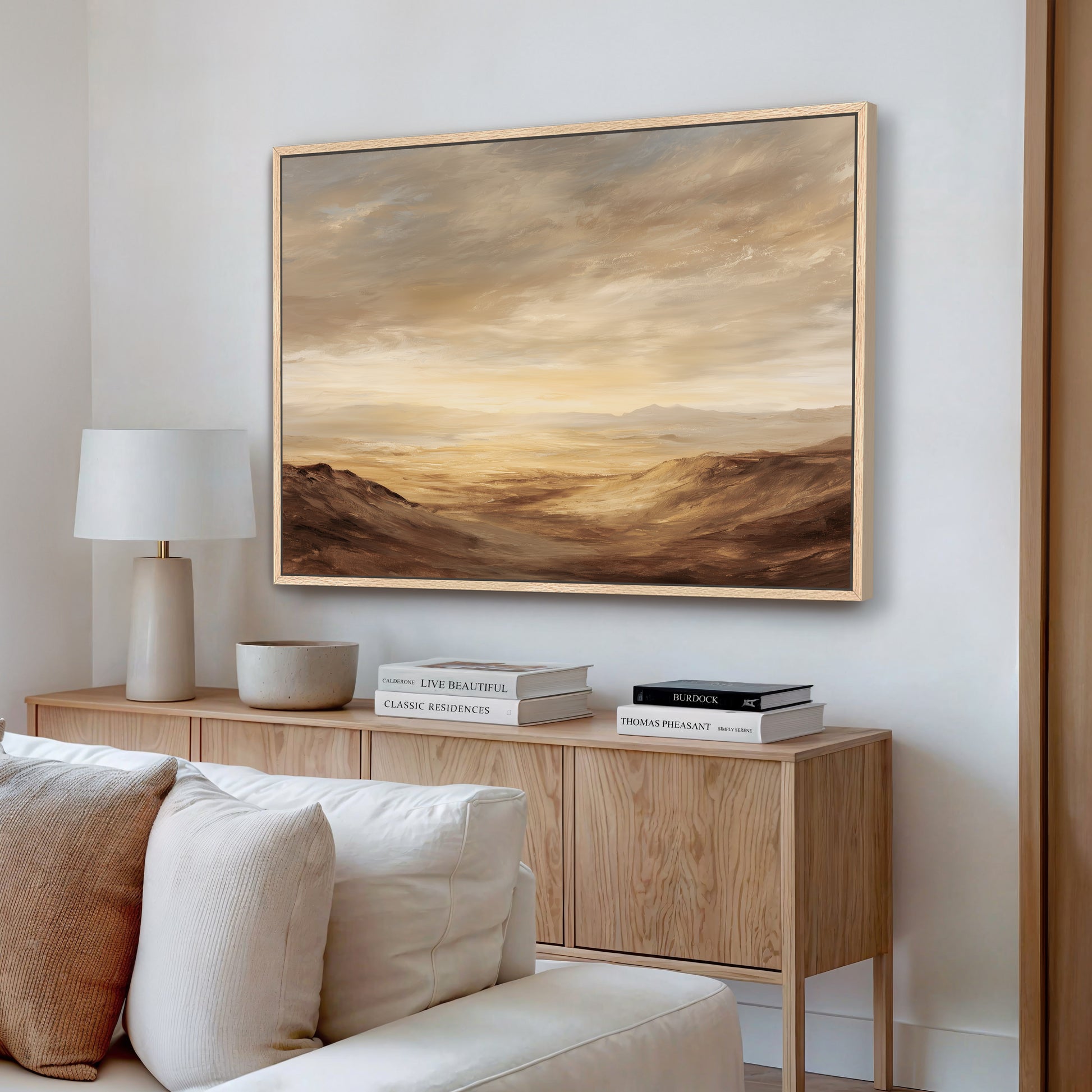 a living room with a large painting of a desert landscape hanging on the wall above a wooden cabinet. The cabinet has a lamp and some books on it. The room has a white couch and a wooden side table.