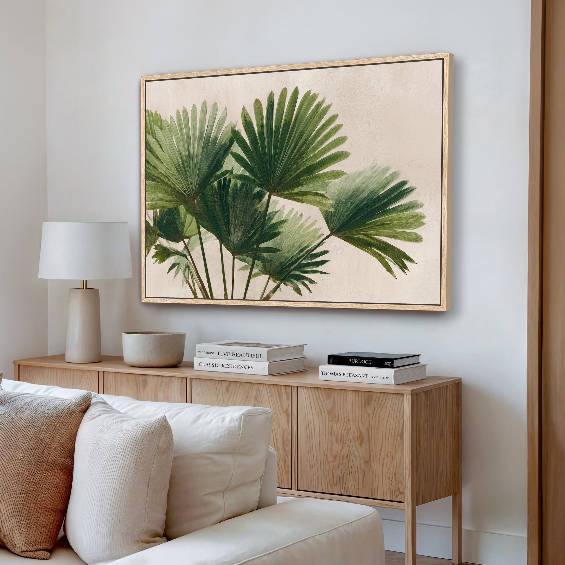 a living room with a large framed painting of green palm leaves hanging on the wall above a wooden cabinet. The cabinet has a bowl and a stack of books on top. There is a white couch in the room, and the overall color scheme is neutral, with the green leaves of the painting adding a touch of nature to the space.