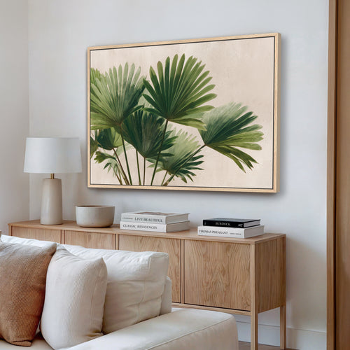 a living room with a large framed painting of green palm leaves hanging on the wall above a wooden cabinet. The cabinet has a bowl and a stack of books on top. There is a white couch in the room, and the overall color scheme is neutral, with the green leaves of the painting adding a touch of nature to the space.