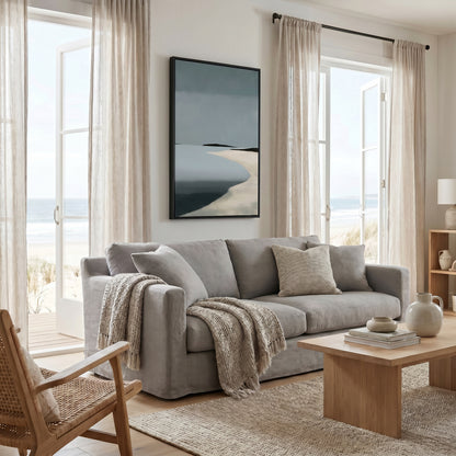 a cozy living room with a large window overlooking the ocean, featuring a gray sofa, a wooden coffee table, and a framed artwork on the wall.