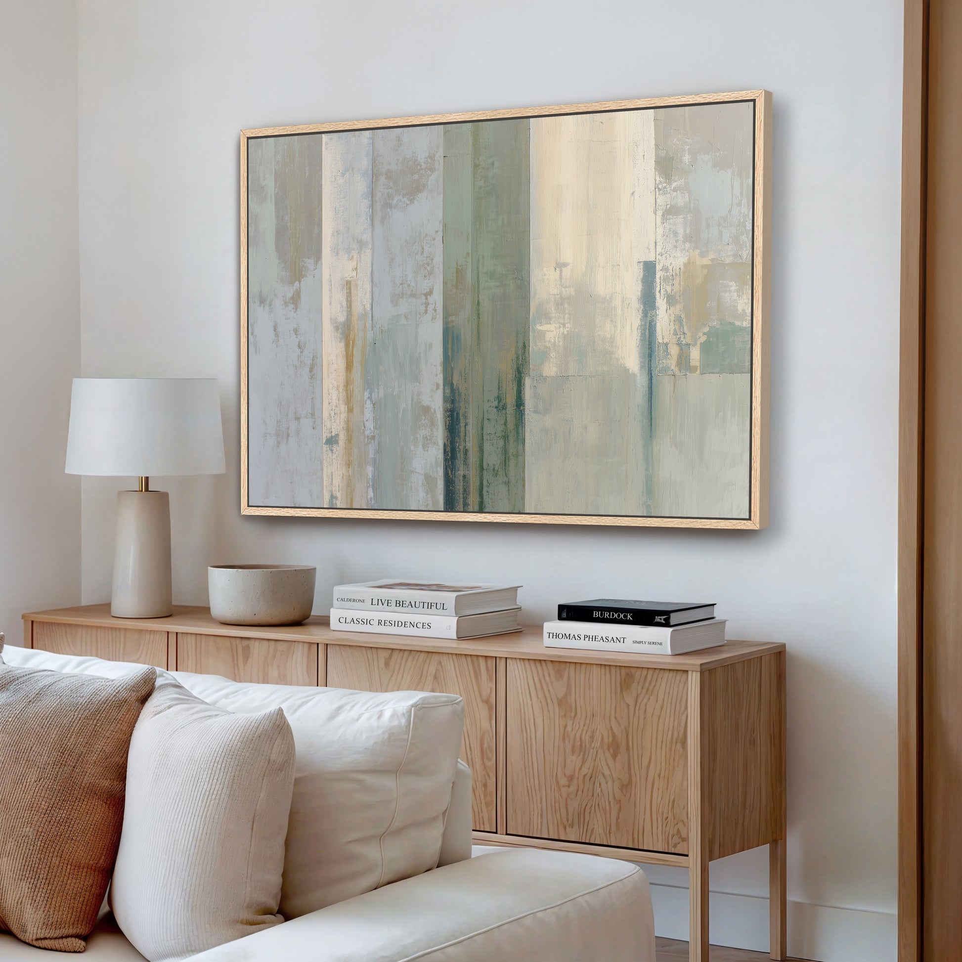 a modern living room with a large abstract painting hanging on the wall above a wooden console table. The console table has a lamp and a stack of books on it. The room also includes a white sofa and a wooden bookshelf.