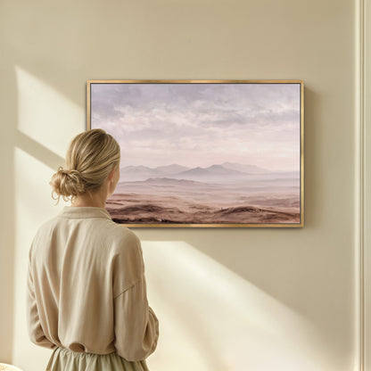 A woman stands in front of a framed landscape painting, gazing at the serene scene of mountains and a hazy sky.