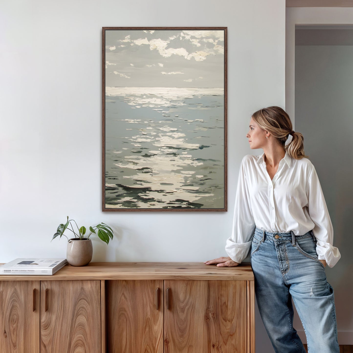 A woman in a white blouse and blue jeans stands next to a wooden cabinet, leaning against it and looking to the side. Above her, a framed painting of a serene ocean scene hangs on the wall.