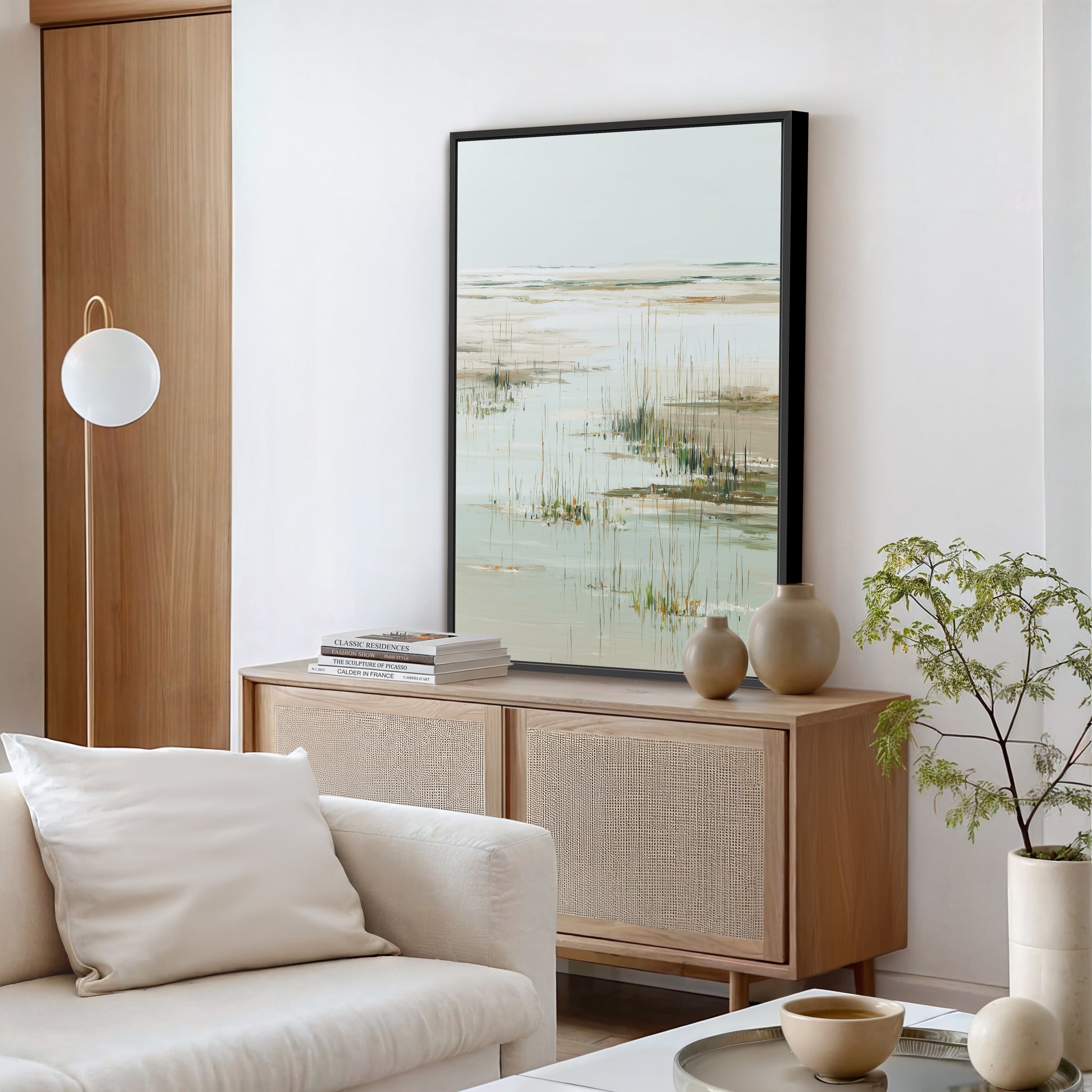 a modern living room with a large painting of a serene landscape hanging on the wall above a wooden cabinet. The cabinet has two vases and a stack of books on top. The room also features a white sofa, a potted plant, and a bowl on a coffee table.