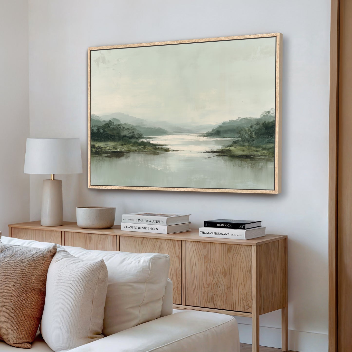a living room with a large landscape painting hanging on the wall above a wooden cabinet. The painting depicts a serene scene of a lake surrounded by mountains. The cabinet has a white lamp and a stack of books on top. The room has a white sofa and a wooden coffee table, creating a cozy and inviting atmosphere.