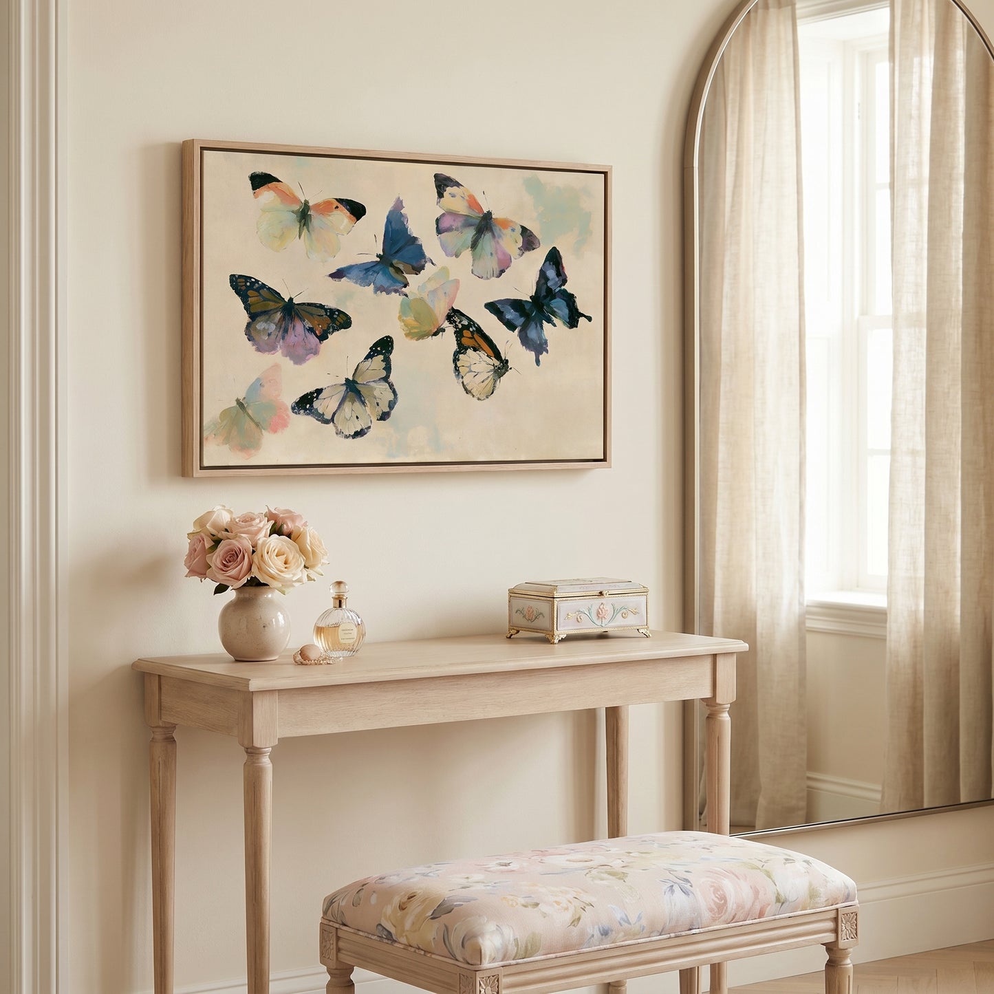 a light-colored wooden table with a vase of flowers, a small box, and a mirror above it. The table is situated in a room with a window and a painting of colorful butterflies hanging on the wall.