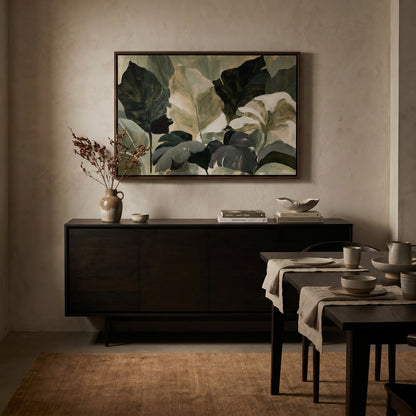 a dining room with a large painting of green leaves hanging on the wall above a dark wooden cabinet. The cabinet has a vase of dried flowers and a bowl on top. In front of the cabinet, there is a table set with plates, bowls, and a vase of flowers. The room has a neutral color palette with a brown rug on the floor.