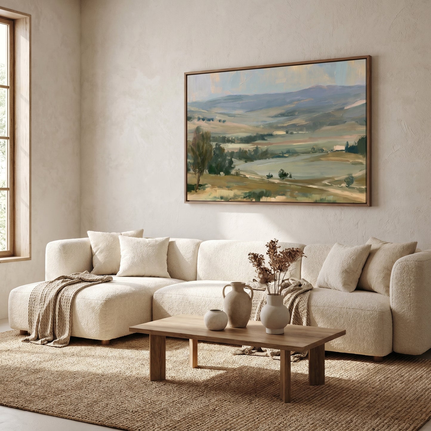 a cozy living room with a large painting of a countryside landscape hanging on the wall above a white sofa. The sofa is positioned in front of a wooden coffee table, which has a vase of flowers and a small white pitcher on it. The room has a neutral color scheme with beige walls and a brown rug, creating a warm and inviting atmosphere.
