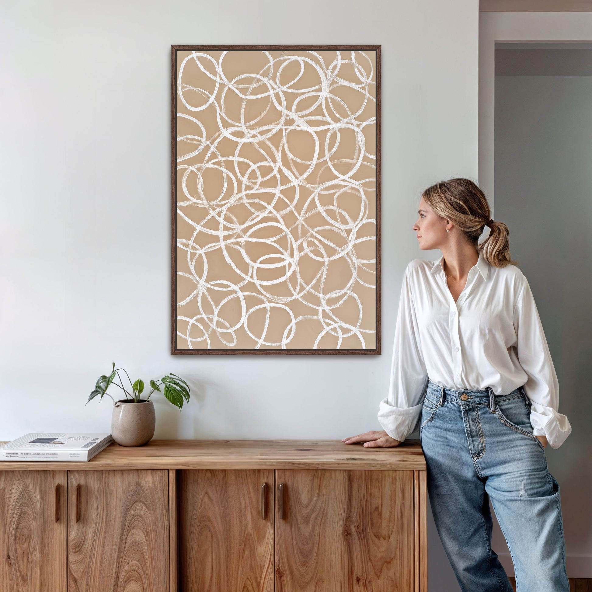 A woman in a white shirt and blue jeans stands next to a wooden cabinet, leaning against it with her hand on her hip. Above her, a framed abstract artwork with a circular pattern hangs on the wall.