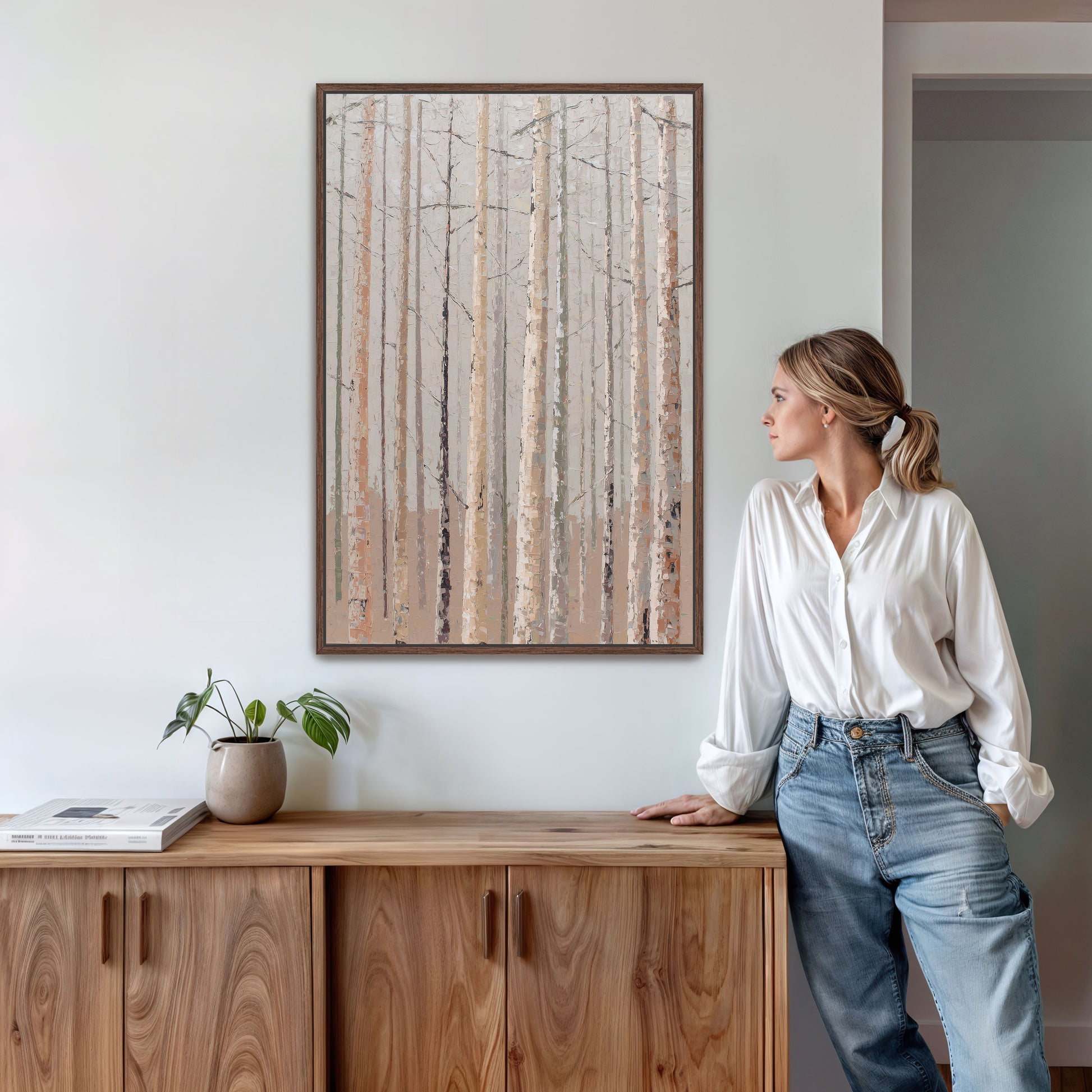 A woman in a white blouse and blue jeans stands in a room with a wooden cabinet, a potted plant, and a framed painting of a forest on the wall.