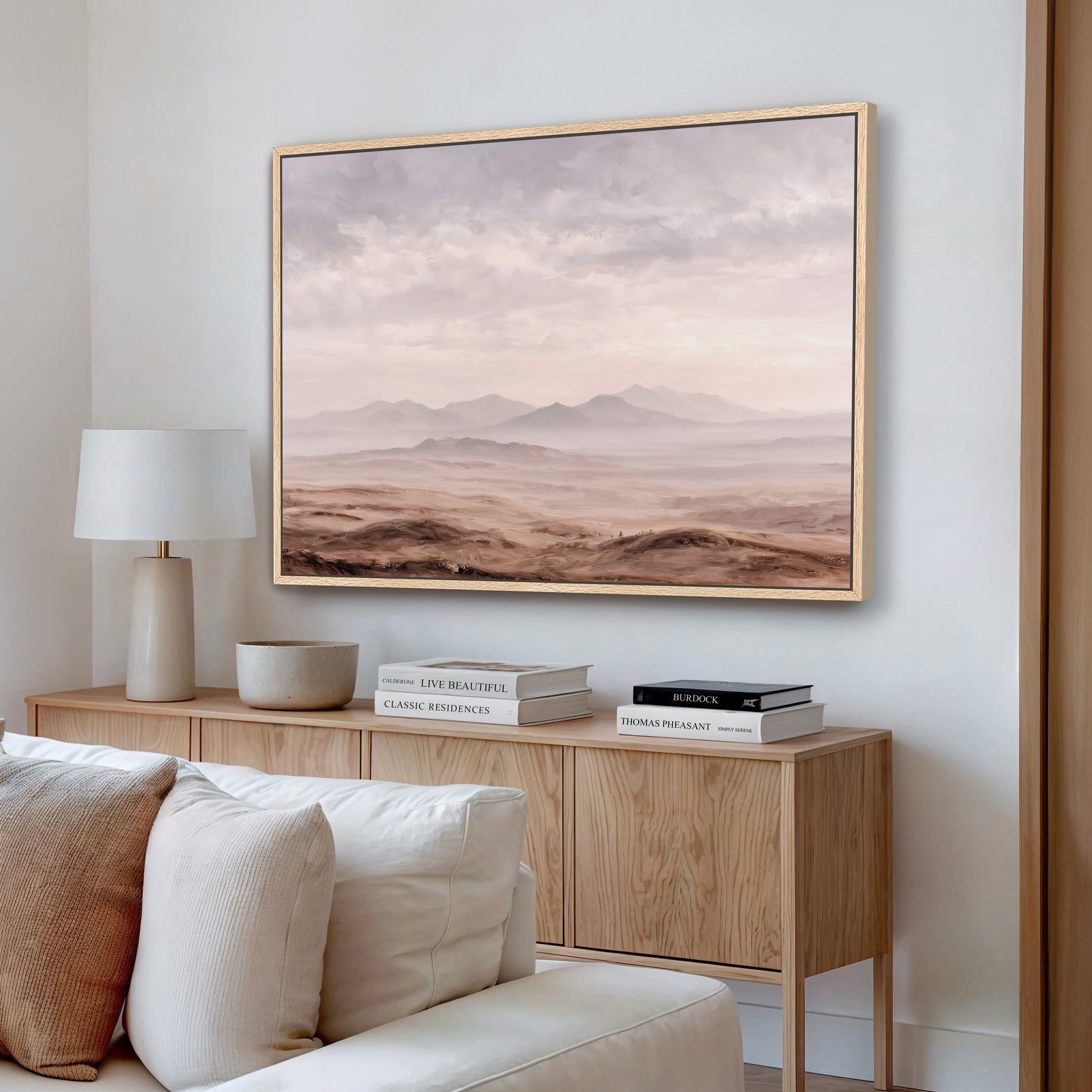 a living room with a large framed landscape painting hanging on the wall above a wooden cabinet. The painting depicts a desert scene with mountains in the background. The cabinet has a lamp and some books on it, creating a cozy and inviting atmosphere.