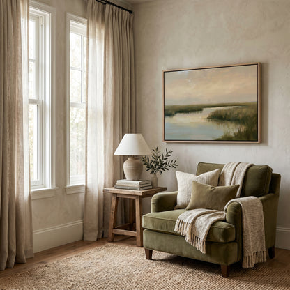 a cozy living room with a large window, a painting of a serene landscape, a wooden side table with a lamp and a plant, and a green armchair with a throw blanket draped over it.