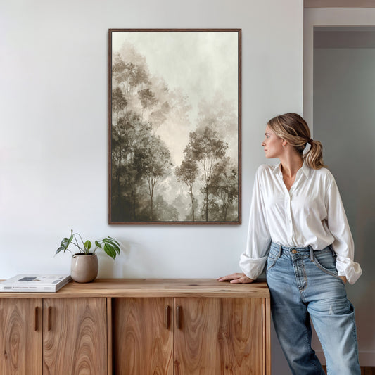 A woman in a white blouse and blue jeans stands in a room, leaning against a wooden cabinet, with a framed landscape painting hanging on the wall above her.