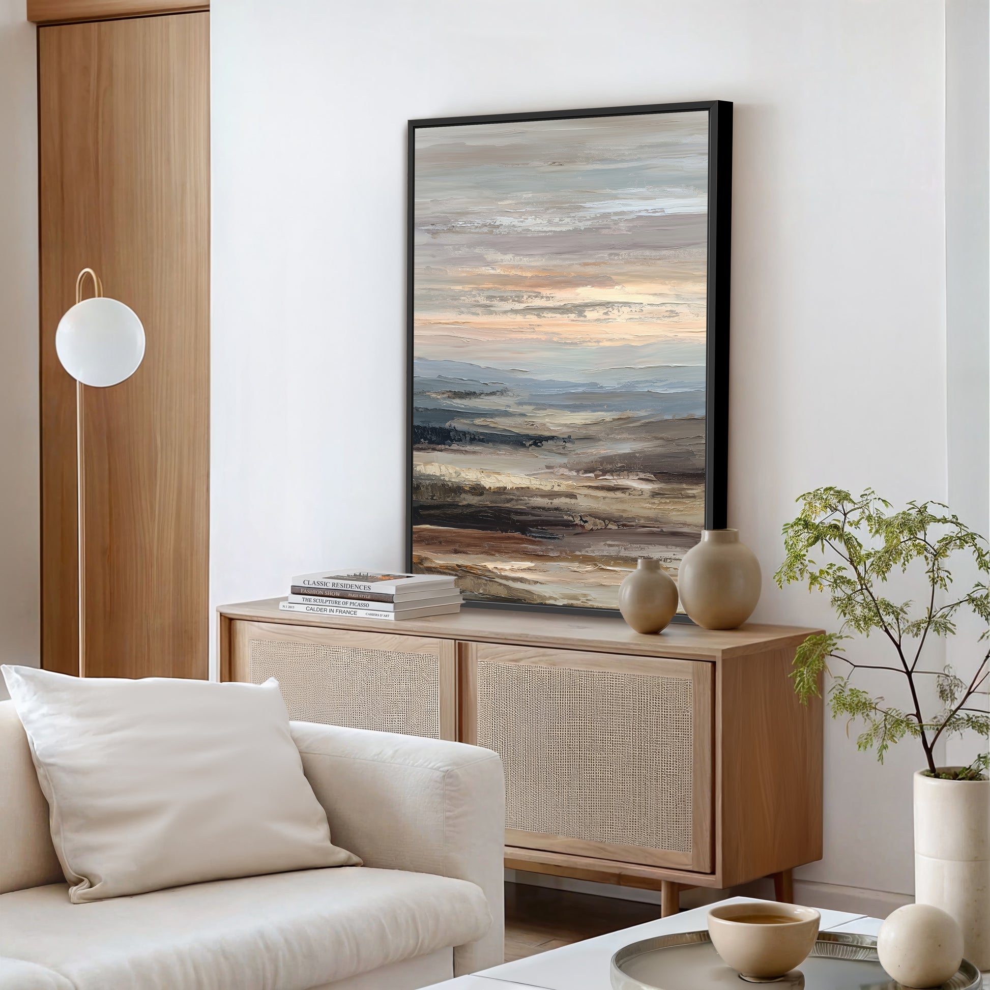 a modern living room with a large painting of a landscape hanging on the wall above a wooden cabinet. The cabinet has two vases and a stack of books on top. The room also features a white sofa, a potted plant, and a round table with a bowl and a plant on it.