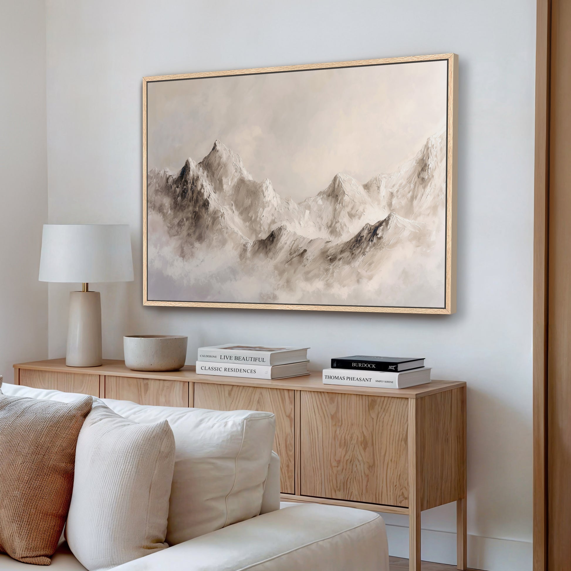 a living room with a large framed painting of a mountain landscape hanging on the wall above a wooden cabinet. The cabinet has a bowl and a stack of books on top. The room also includes a white couch and a lamp on a side table.