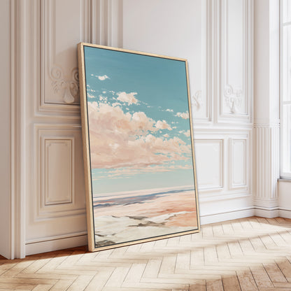 A large framed painting depicting a serene landscape with a blue sky, fluffy clouds, and a sandy beach is leaning against a white wall in a room with a wooden floor.