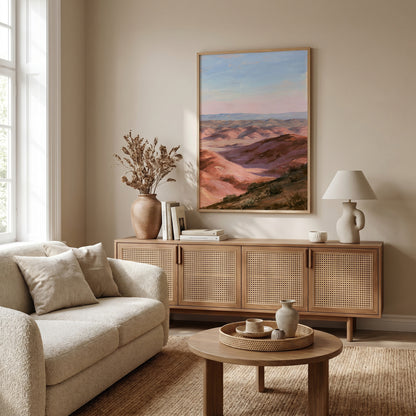 a cozy living room with a large painting of a mountainous landscape hanging on the wall. The room is furnished with a beige sofa, a wooden sideboard, and a round coffee table. The room is decorated with various items, including a vase, a lamp, and a book, creating a warm and inviting atmosphere.