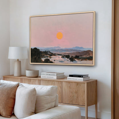 a living room with a large painting of a sunset landscape hanging on the wall above a wooden cabinet. The cabinet has a bowl and a stack of books on it. The room also includes a white couch and a lamp.