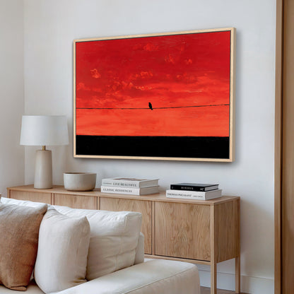 a living room with a large framed painting of a red sky with a bird on a wire hanging on the wall above a wooden cabinet. The cabinet has a white bowl and a stack of books on top. The room also includes a white couch and a lamp.