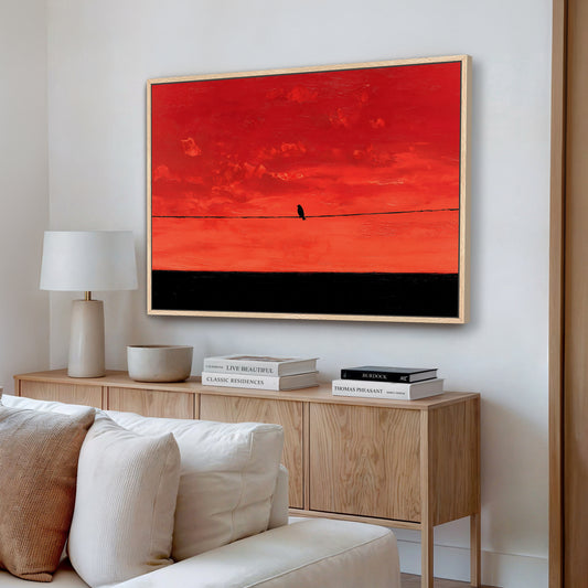 a living room with a large framed painting of a red sky with a bird on a wire hanging on the wall above a wooden cabinet. The cabinet has a white bowl and a stack of books on top. The room also includes a white couch and a lamp.
