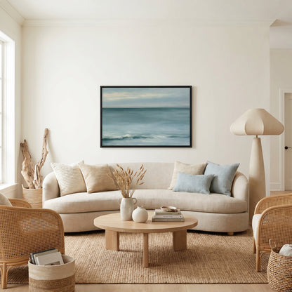 a serene living room with a large painting of a calm ocean scene hanging on the wall. The room is furnished with a beige sofa, a wooden coffee table, and a woven chair. The overall decor has a minimalist and coastal theme, with natural elements such as a vase of dried flowers and a potted plant adding to the ambiance.