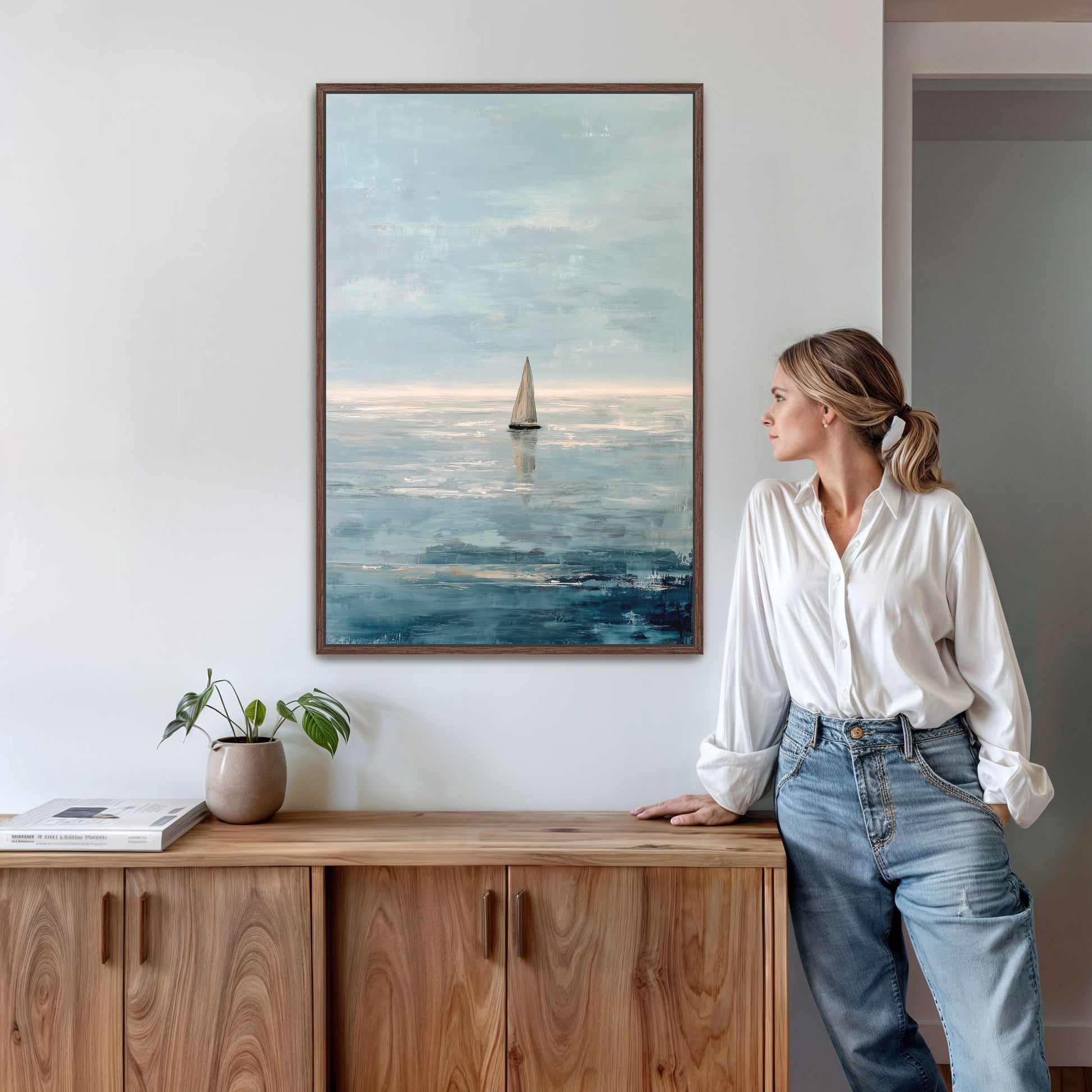 A woman in a white blouse and blue jeans stands in a room, leaning against a wooden cabinet, with a framed painting of a sailboat on the wall behind her.