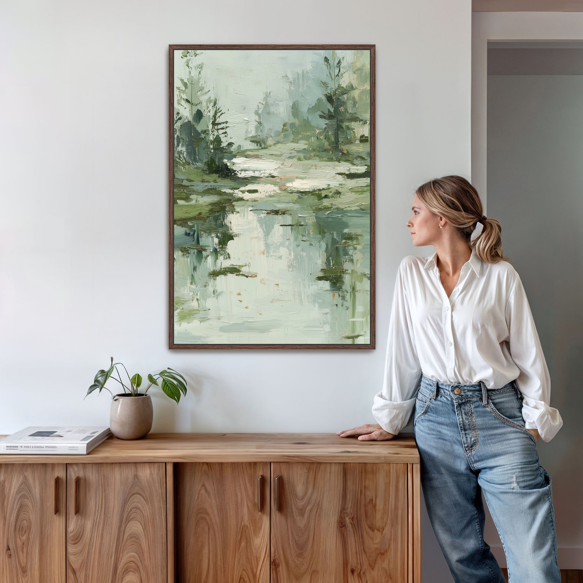 A woman stands in a room, leaning against a wooden cabinet, with a framed landscape painting hanging on the wall above her.