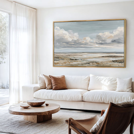 a serene living room with a large painting of a cloudy sky hanging on the wall above a white sofa. The room is furnished with a wooden coffee table, a brown armchair, and a wooden side table. The overall decor has a minimalist and modern aesthetic.