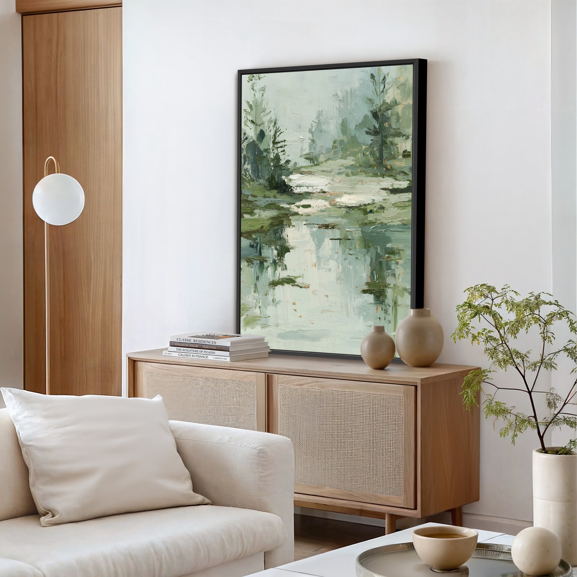 a serene living room with a large painting of a forest scene hanging on the wall above a wooden cabinet. The cabinet has two vases and a stack of books on top. The room also features a white sofa, a potted plant, and a bowl on a coffee table.