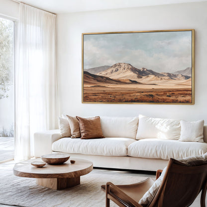 a living room with a large painting of a desert landscape hanging on the wall above a white sofa. The sofa is positioned in front of a wooden coffee table, which has a bowl and a vase on it. There is also a chair in the room. The overall decor has a minimalist and modern style, with a focus on neutral colors and natural elements.