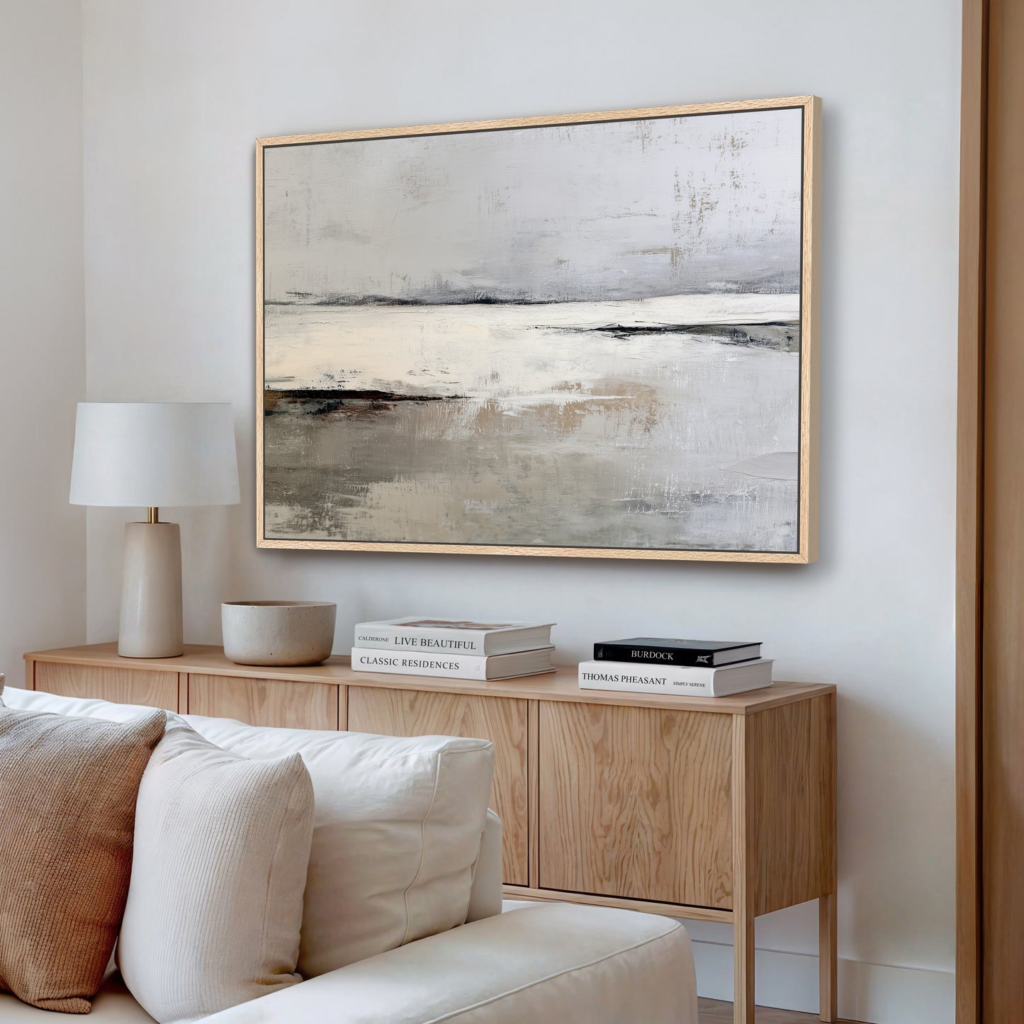 a living room with a large abstract painting hanging on the wall above a wooden cabinet. The cabinet has a lamp and some books on it. The painting has a muted color palette and depicts a landscape scene.