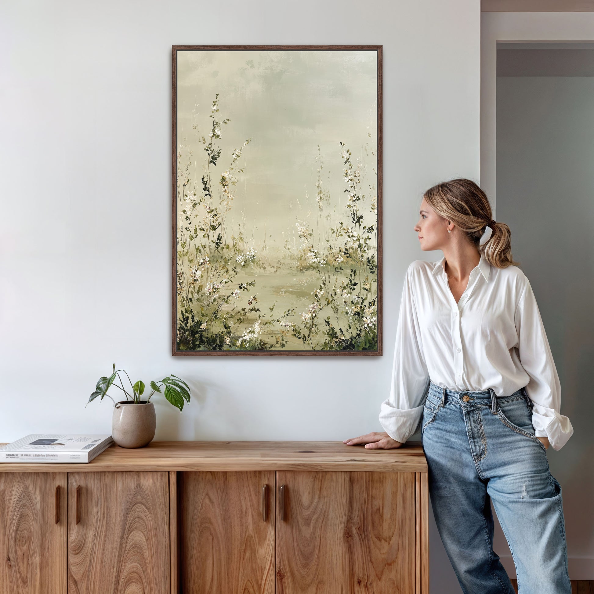 A woman in a white blouse and blue jeans stands next to a wooden cabinet, leaning against it and looking to the side. Above her, a framed painting of flowers hangs on the wall.