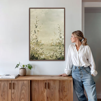 A woman in a white blouse and blue jeans stands next to a wooden cabinet, leaning against it and looking to the side. Above her, a framed painting of flowers hangs on the wall.