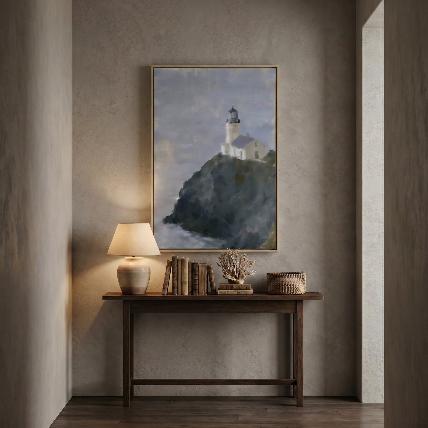 a wooden console table with a lamp and a stack of books, set against a wall with a framed painting of a lighthouse on a cliff.