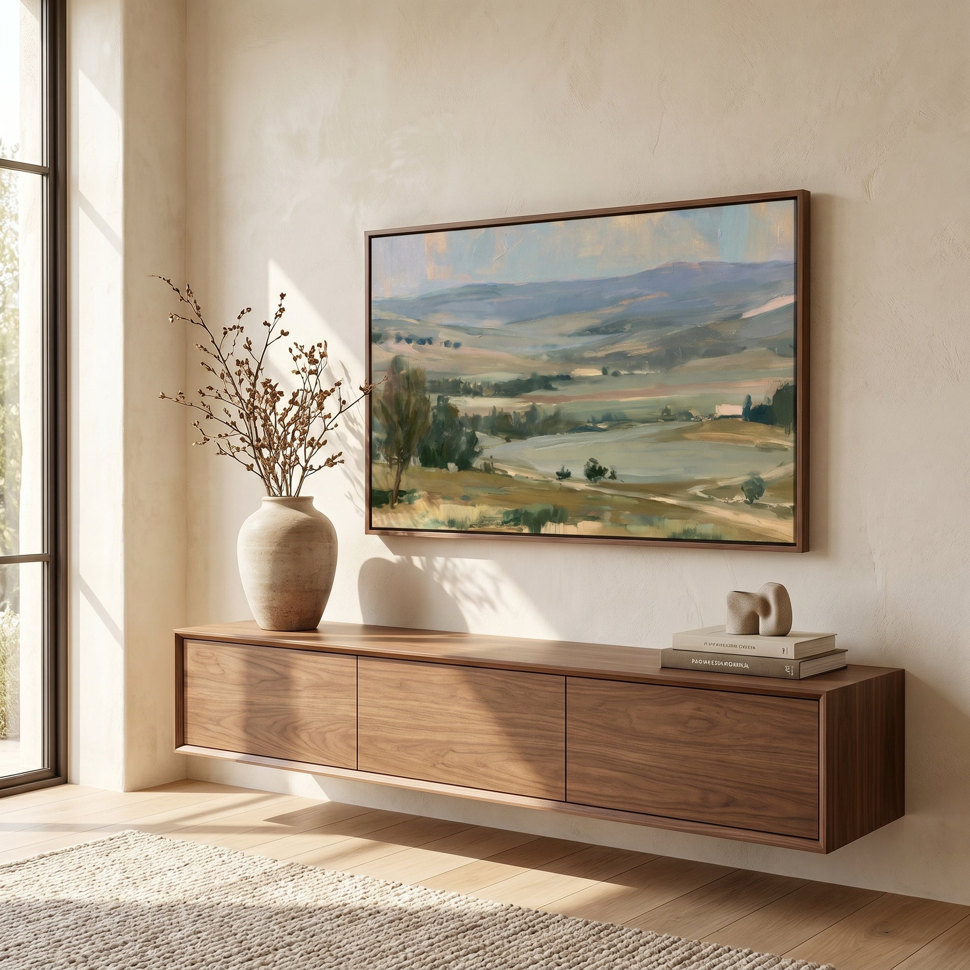 a large landscape painting hanging on a wall above a wooden cabinet or shelf. The painting depicts a serene countryside scene with rolling hills, trees, and a winding road. The cabinet or shelf below the painting has a vase and some books placed on it, adding a touch of personalization to the space.