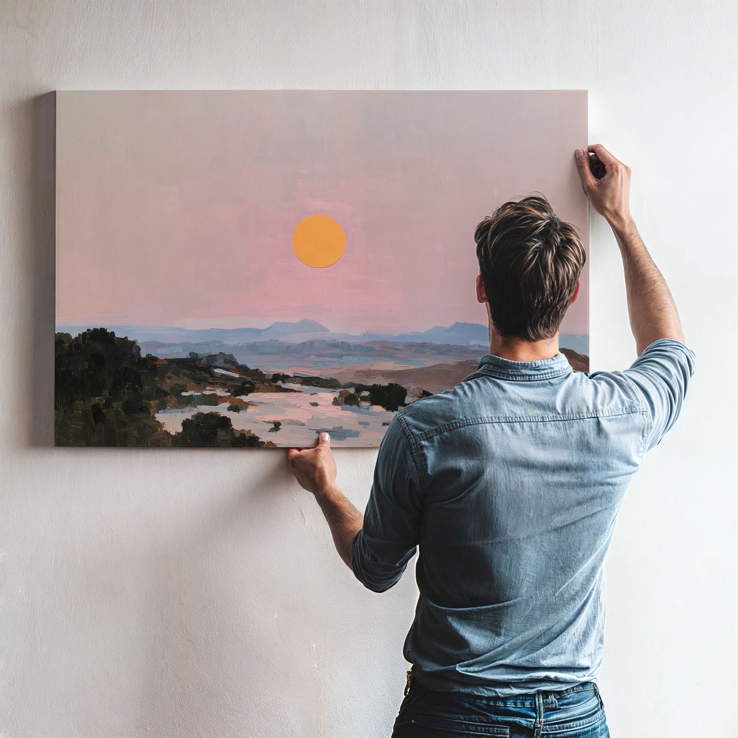 A man is hanging a large landscape painting on a wall, which depicts a serene sunset over a mountainous terrain.